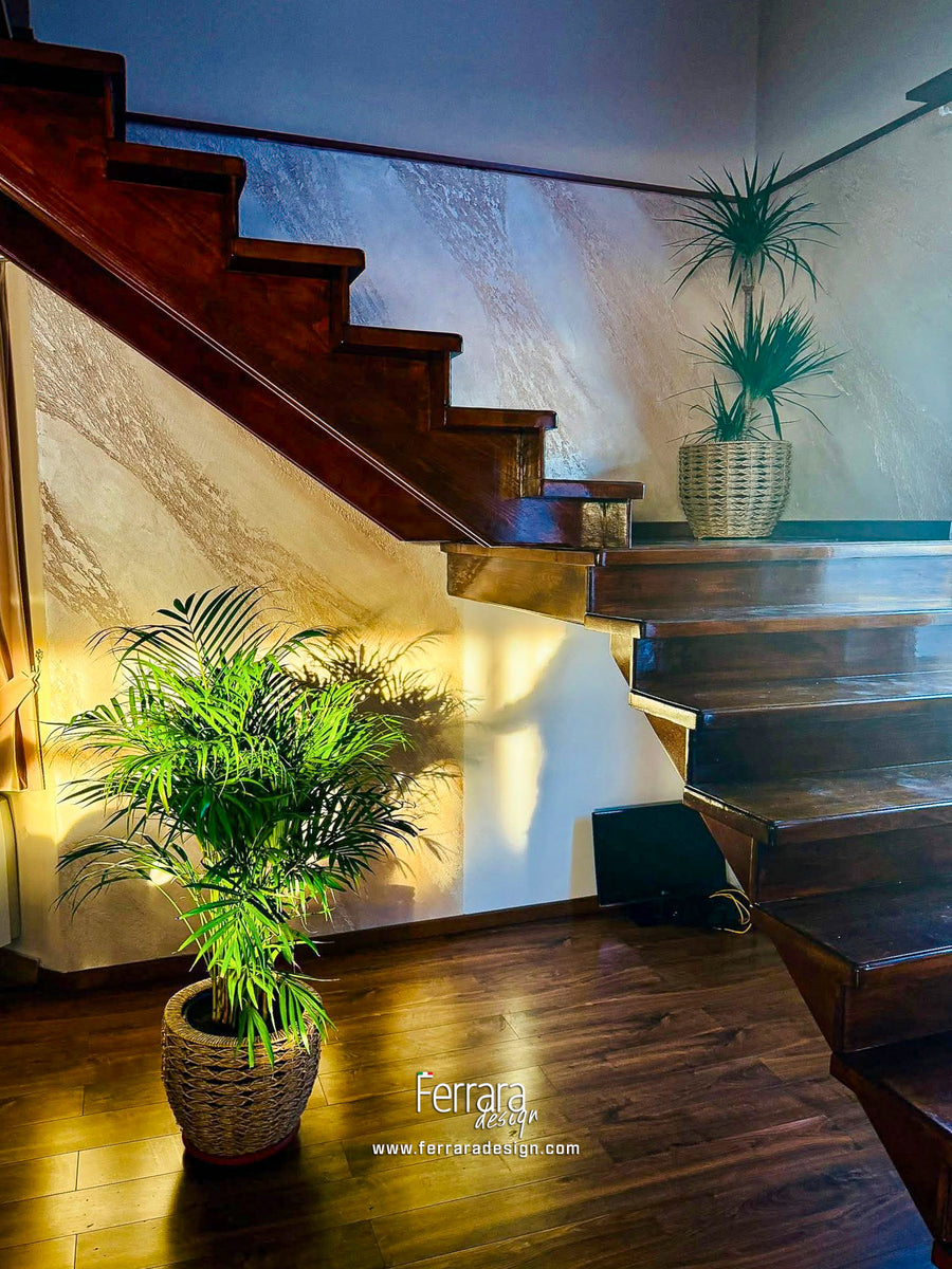 A modern staircase with dark wood steps and a light stone wall, decorated with potted green plants in woven baskets. The word 'Ferrara' and a website URL are visible at the bottom.