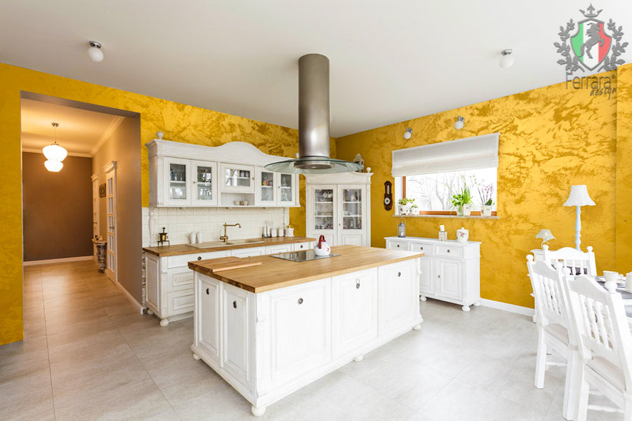 A bright kitchen interior with white cabinetry and island, light wood countertops, a tall stainless steel range hood, textured yellow walls, tiled flooring, and a dining chair visible at the right. A watermark/logo appears in the top-right corner.