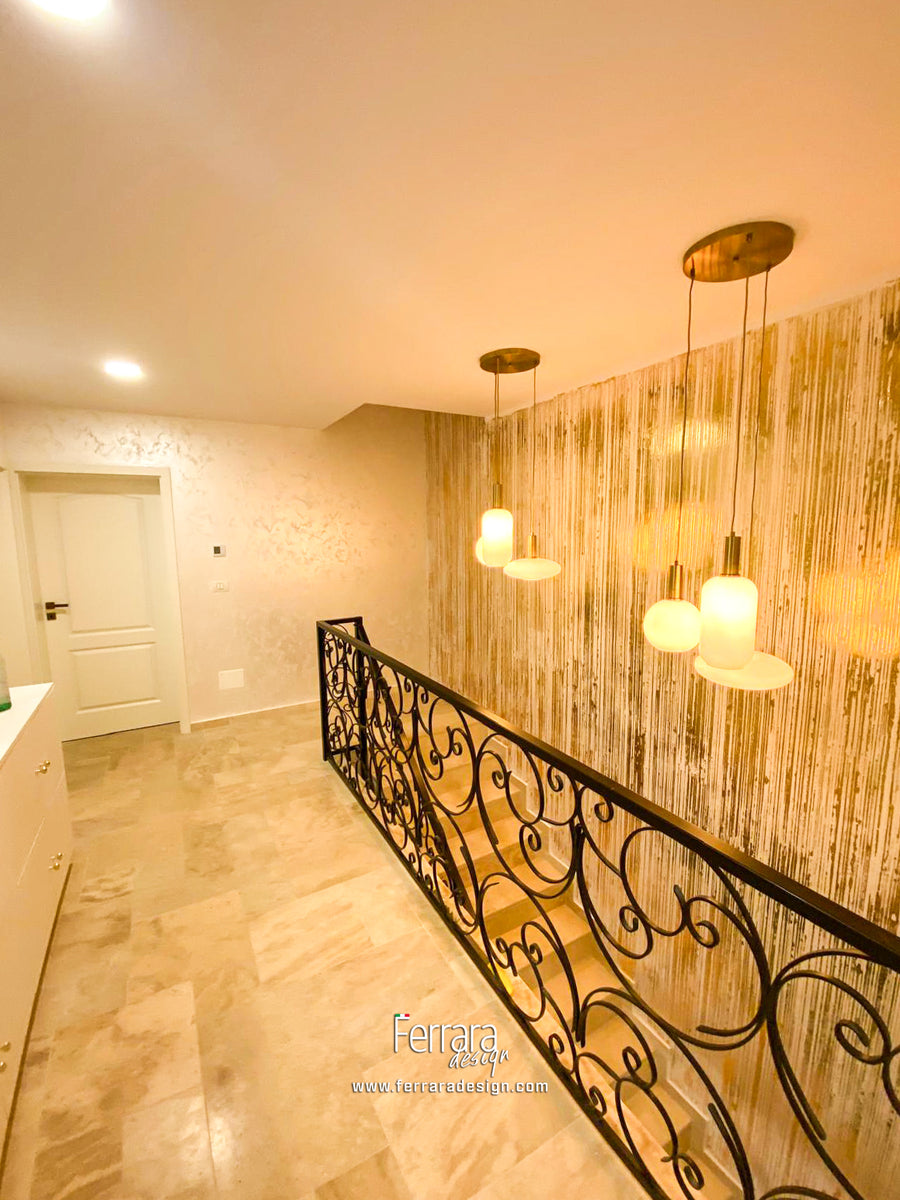 An interior staircase landing with a black wrought-iron scroll railing, warm beige walls, a wood-look accent wall, and three hanging pendant lights with frosted white shades.