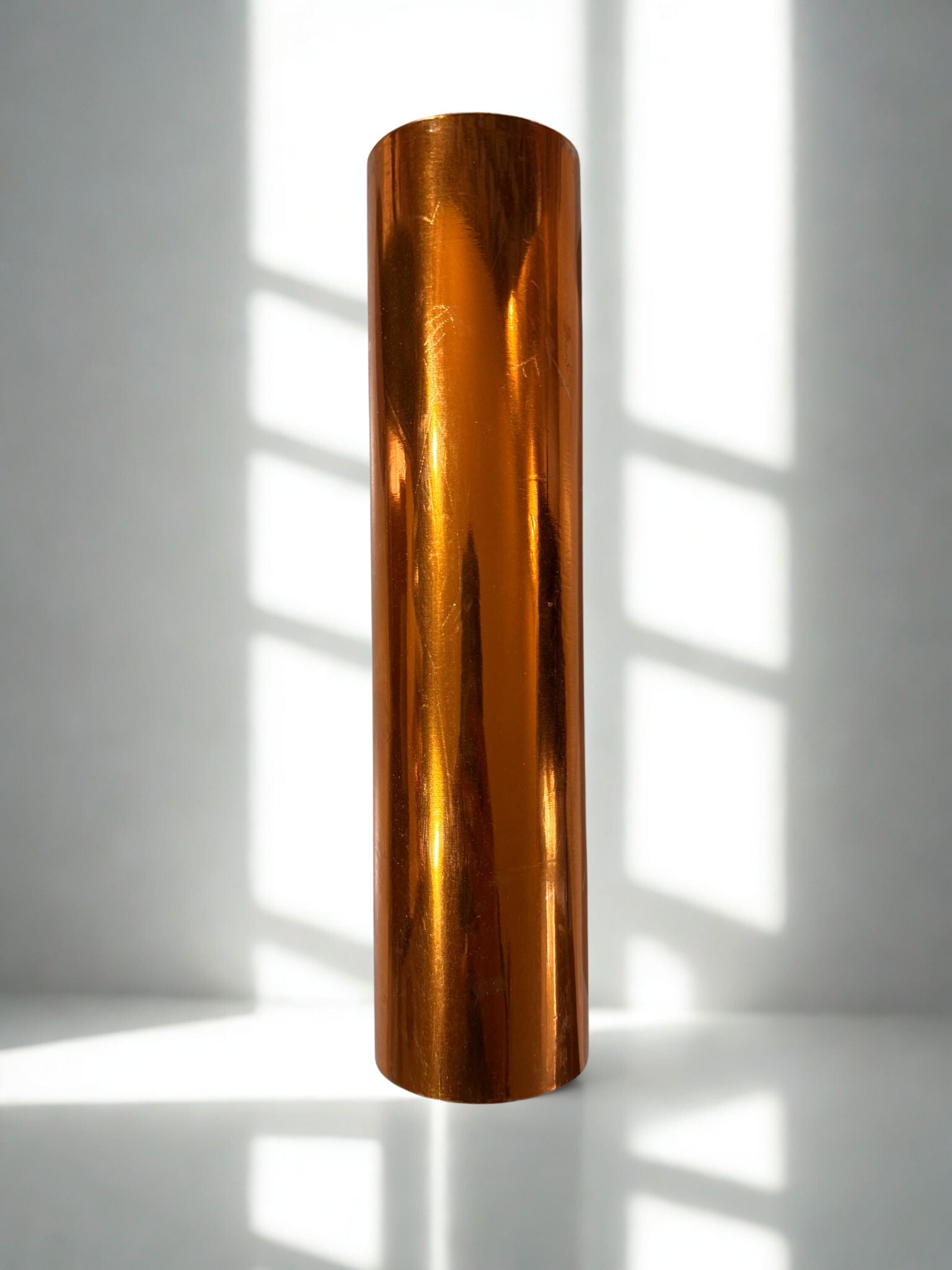 Bronze-colored cylindrical object on a white surface with shadows from vertical blinds.
