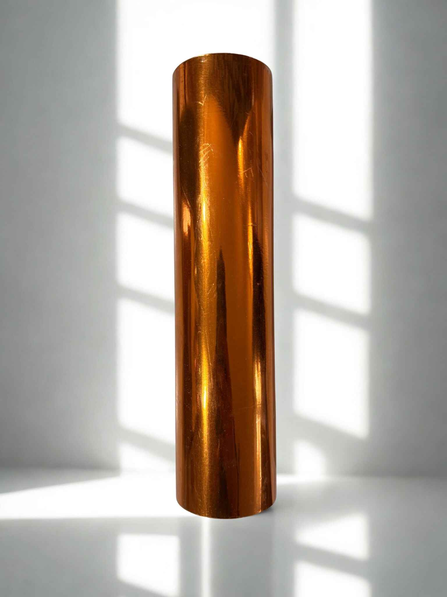 Bronze-colored cylindrical object on a white surface with shadows from vertical blinds.